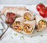 Stack of veggie burritos on a place next to a glass pot of chopped tomatoes