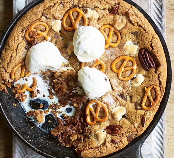 A giant cookie in a skillet topped with marshmallows, pretzels and nuts